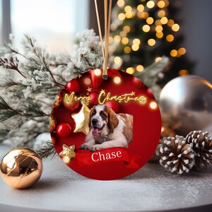 May include: A round Christmas ornament with a photo of a dog named Chase. The red ornament has gold text that says "Merry Christmas" and is decorated with gold stars and red ornaments. A gold ribbon is attached for hanging.