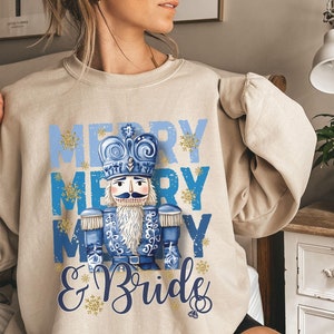 May include: A beige sweatshirt with a blue and white design featuring a nutcracker and the text "Merry Merry Merry & Bride".