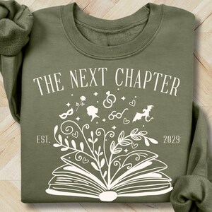 May include: Military green sweatshirt with the text "THE NEXT CHAPTER" and an open book design. The design includes floral elements, a mask, and the year 2029. The sweatshirt is a crew neck style.