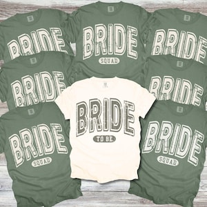 May include: Set of six t-shirts with the word "Bride" printed on them in a distressed font. One shirt says "Bride to Be" and the others say "Bride Squad". The shirts are in two colors: ivory and moss green.