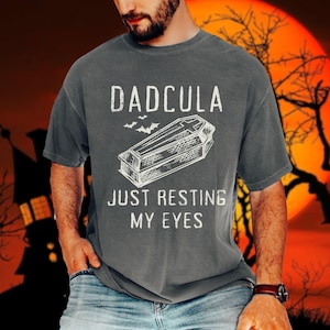 May include: A gray t-shirt with the text "DADCULA JUST RESTING MY EYES" in white. A black and white illustration of a coffin with a cross on top is above the text. There are three bats flying above the coffin.