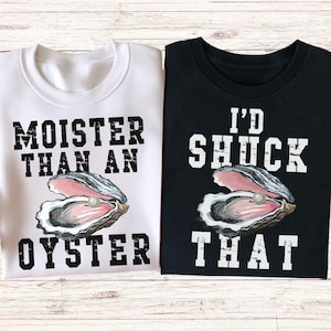 May include: Two t-shirts, one white and one black, with oyster-themed graphics. The white shirt says "Moister Than An Oyster" and the black shirt says "I'd Shuck That". Each shirt features an illustration of an open oyster with a pearl.