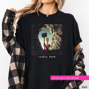 May include: Black t-shirt with a square graphic featuring a doll holding a shovel, wearing a "Bride" sash, and emerging from a hole. The words "custom text" are below the graphic. A pink banner at the bottom says "Choose your own phrase".