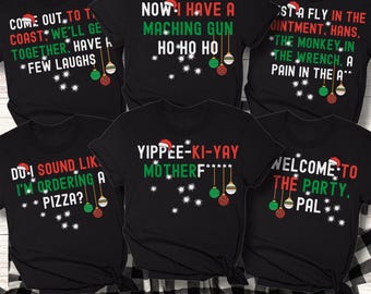 May include: Black t-shirts with humorous Christmas-themed phrases in white and green text. Each shirt features a Santa hat and hanging ornaments. Phrases include "Now I Have A Maching Gun Ho Ho Ho" and "Welcome to the Party, Pal."