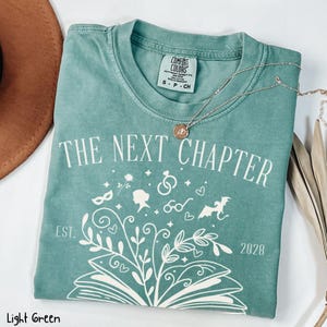 May include: Light green t-shirt with the text "THE NEXT CHAPTER" and a graphic of a book with floral and whimsical elements. The shirt also features the text "EST. 2028". A silver necklace with a pendant rests on the shirt.