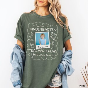 May include: A moss green t-shirt with white text and a photo of a child. The text reads "Forever in Kindergarten, Teacher Crew, best team ever" with the name "Mrs. Johnson" below the photo. The shirt is a relaxed fit.