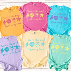 May include: A collection of six colorful t-shirts. Each shirt features the text "Toes in the sand" and "Drink in my hand" or "Ring on my hand" with seashell, cocktail, and starfish graphics. Colors include yellow, pink, blue, and purple.