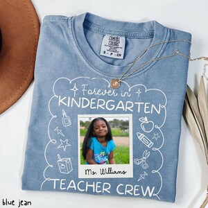 May include: Blue jean colored t-shirt with the text "Forever in Kindergarten TEACHER CREW". The shirt features a photo of a young girl and the name "Ms. Williams". A gold necklace is draped across the shirt.
