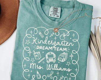 May include: Light green t-shirt with the text "Kindergarten DREAM TEAM Mrs. Williams" in white, featuring school-themed illustrations. A silver necklace with a pendant is draped across the shirt.