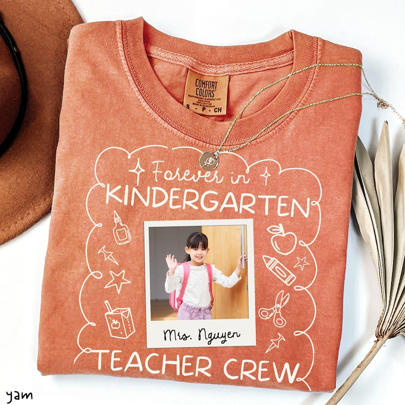 May include: A coral-colored crew neck sweatshirt with the words "Forever in Kindergarten TEACHER CREW" in white script. A photo of a young girl with a backpack is in the center. A gold necklace is draped across the shirt.