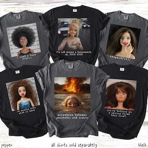 May include: A collection of black and gray graphic t-shirts. Each shirt features a doll image with text. The text includes phrases like "I need a huge cock...", "I'm not always a trainwreck", and "somewhere between psychotic and iconic."