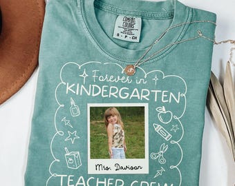 以下が含まれることがあります： 「Forever in Kindergarten TEACHER CREW」の文字が入った、ライトグリーンのComfort Colors Tシャツ。中央には少女の写真が配置されています。シルバーのネックレスがシャツの上にかけられています。