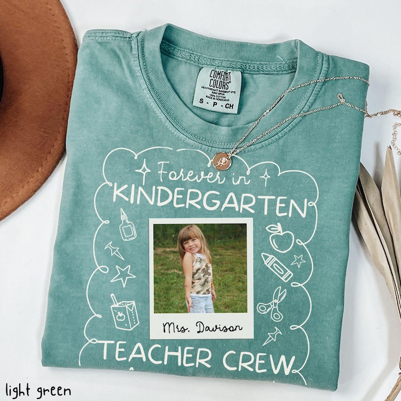 May include: Light green Comfort Colors t-shirt with the text "Forever in Kindergarten TEACHER CREW". A photo of a young girl is in the center. A silver necklace is draped across the shirt.