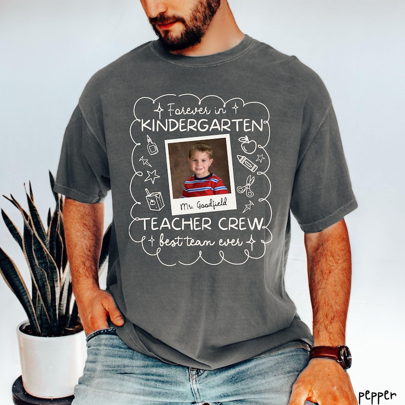 May include: Dark gray t-shirt with a framed photo of a child and the words "Forever in Kindergarten, Teacher Crew, best team ever." The photo is surrounded by school-themed doodles. The shirt is a comfortable, casual style.