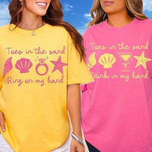 May include: Two short-sleeved t-shirts, one yellow and one pink, with beach-themed graphics and text. The yellow shirt says "Toes in the sand, Ring on my hand" with seashell, ring, and starfish icons. The pink shirt says "Toes in the sand, Drink in my hand" with seashell, cocktail, and starfish icons.