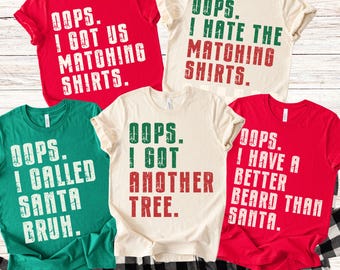 May include: A collection of five short-sleeved t-shirts in red, green, and cream colors. Each shirt has a humorous holiday-themed phrase printed in bold, white, green, and red text. The shirts are folded on a black and white checkered fabric.