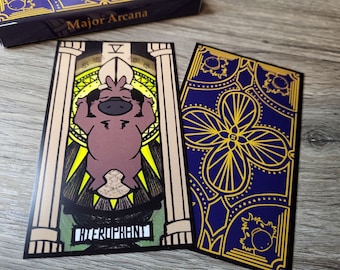 Könnte beinhalten: Ein Kartenspiel der Tarotkarten der Großen Arkana. Eine Karte zeigt einen braunen Cartoon-Charakter mit dem Wort "HIEROPHANT" unten. Die andere Karte hat einen blauen Hintergrund mit einem goldenen Blumendesign. Die Box trägt die Aufschrift "Major Arcana".