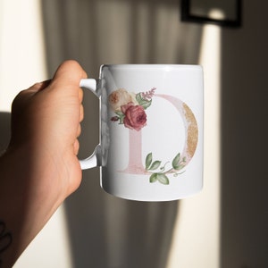 May include: White ceramic mug with a pink and gold letter D decorated with watercolor flowers. The flowers are pink, white, and green.
