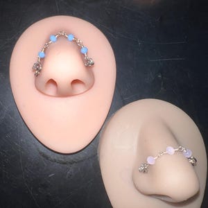 May include: Close-up of two nose jewelry pieces. One features a silver chain with light blue beads and decorative charms. The other has a similar design with pink beads. Both are displayed on a flesh-toned nose form.
