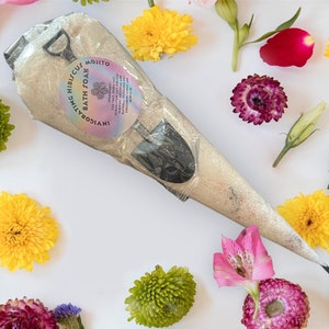 Puede incluir: Una bolsa blanca en forma de cono llena de baño de burbujas. La bolsa tiene una etiqueta que dice "Invigorating Hibiscus Bath Soak Mojito". La bolsa está rodeada de varias flores de diferentes colores.