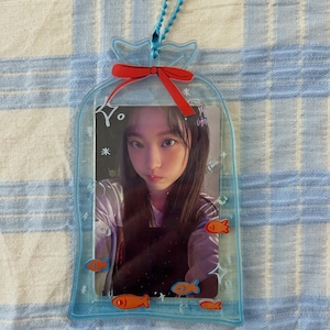 May include: A clear plastic photo frame shaped like a bag with a red bow and a blue chain. The frame has a picture of a young woman inside and orange fish illustrations around the edge.