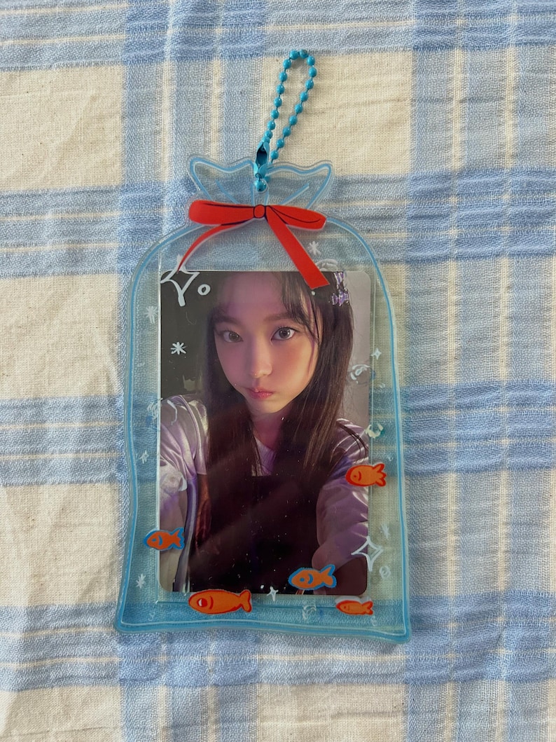May include: A clear plastic photo frame shaped like a bag with a red bow and a blue chain. The frame has a picture of a young woman inside and orange fish illustrations around the edge.