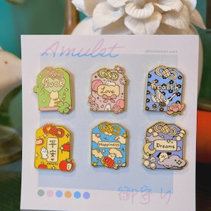 May include: A set of six colorful enamel pins on a white card. Each pin features a unique design with Japanese text and English words like "Luck," "Love," "Health," "Safety," "Happiness," and "Dreams."