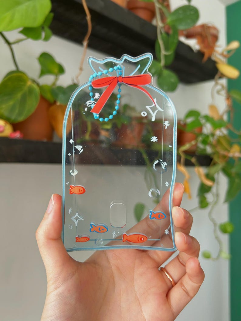 May include: A clear plastic phone case with a blue outline and a red bow. The case has a design of small orange fish and white stars. The case is held in a hand.