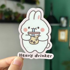 May include: A white bunny rabbit sticker with pink cheeks holding a brown boba tea cup with a blue straw. The sticker has the text "Heavy drinker" below the bunny.