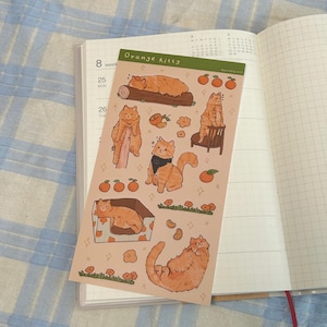 May include: A sheet of stickers featuring orange tabby cats in various poses, including sleeping, sitting, and playing with oranges. The stickers are on a peach-colored background with green grass and orange flowers.