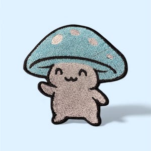 May include: Embroidered patch featuring a smiling mushroom character with a light blue cap and tan body. The mushroom has a black outline and white polka dots on the cap. The patch is designed to be ironed on or sewn onto fabric.