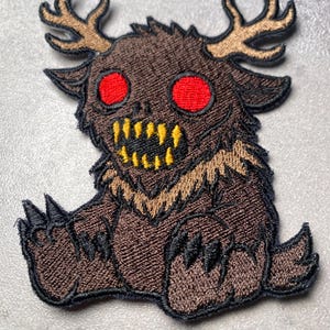 Puede incluir: Parche bordado que representa una criatura de dibujos animados con pelaje marr&oacute;n, cuernos y dientes amarillos. La criatura tiene grandes ojos rojos y garras negras. El parche est&aacute; dise&ntilde;ado con una apariencia detallada y cosida, adecuado para ropa o accesorios.