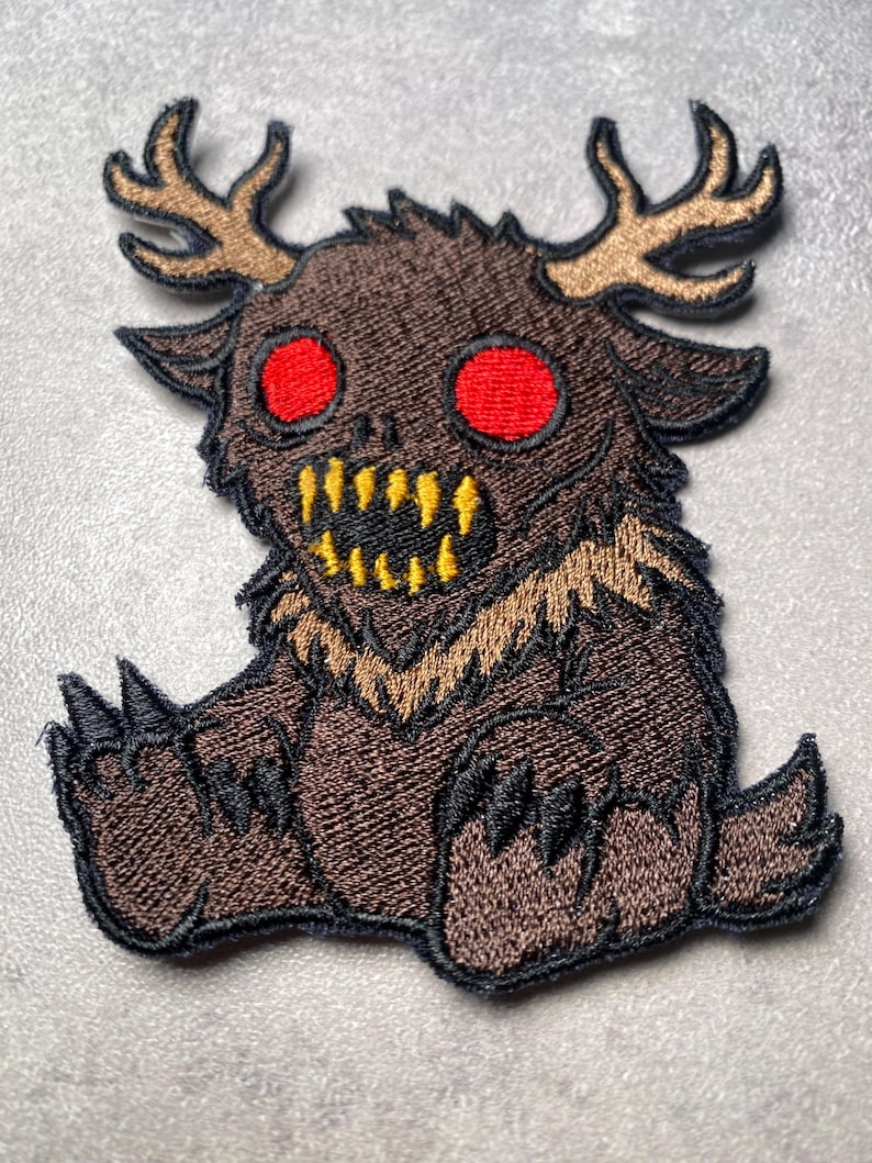 Puede incluir: Parche bordado que representa una criatura de dibujos animados con pelaje marr&oacute;n, cuernos y dientes amarillos. La criatura tiene grandes ojos rojos y garras negras. El parche est&aacute; dise&ntilde;ado con una apariencia detallada y cosida, adecuado para ropa o accesorios.