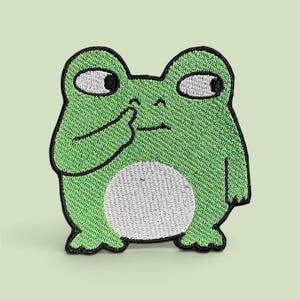May include: An embroidered patch featuring a green frog with a white belly and large white eyes with black pupils. The frog is depicted with one hand raised to its nose, rendered in a cartoon style with a thick black border outlining the entire shape.