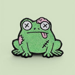 May include: Embroidered patch featuring a green frog with black outlines. The frog has pink patches, crossed-out eyes, and a tongue sticking out. The patch has a stitched appearance, with a black border and cross stitches.