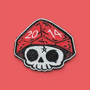Op de afbeelding: Een geborduurde patch met een witte schedel met zwarte oogkassen en een neusholte, bekroond met een rode, gefacetteerde, dobbelsteenvormige pet. De rode pet toont de wit geborduurde cijfers 20 en 14 op de bovenzijde met zwarte omlijningen.