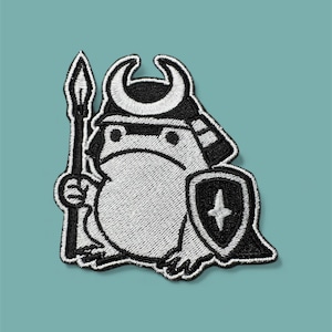May include: Embroidered patch of a cartoon frog in a samurai helmet, holding a spear and shield. The patch is mainly white with black outlines and details. The shield has a white cross design. The background is a solid teal colour.