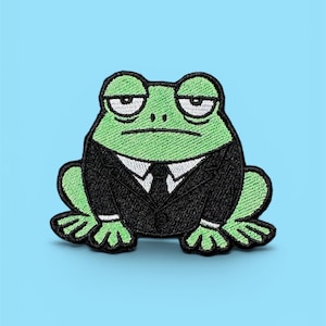 Peut inclure: Écusson brodé représentant une grenouille verte portant un costume noir avec une chemise blanche et une cravate. La grenouille a une expression renfrognée. L'écusson est posé sur un fond bleu clair.