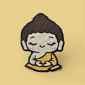 May include: Embroidered patch featuring a cartoon Buddha in a seated meditation pose. The patch has a brown head, white face, pink cheeks, and a yellow robe. The background is a solid light yellow.