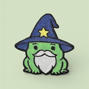 Könnte beinhalten: Gestickter Aufnäher mit einem grünen Frosch, der einen blauen Zaubererhut mit einem gelben Stern trägt. Der Frosch hat einen weißen Bart und einen schwarzen Umriss. Ideal zum Aufnähen auf Kleidung oder Accessoires.
