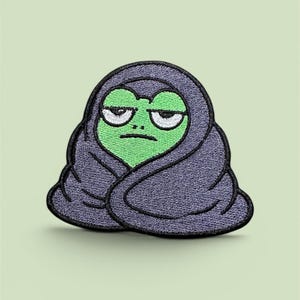 Könnte beinhalten: Ein gestickter Aufnäher, der einen grünen Frosch mit mürrischem Gesichtsausdruck zeigt, eingewickelt in eine dunkelviolette Decke. Der Frosch hat große weiße Augen mit schwarzen Pupillen und einen geraden Mund, alles mit dickem schwarzem Garn umrandet.