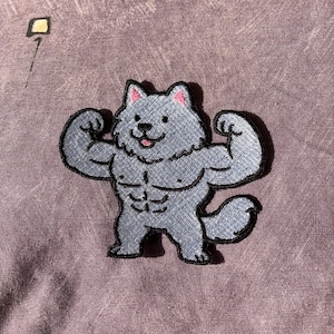 May include: Embroidered patch featuring a cartoon wolf flexing its muscles. The wolf is gray with pink accents on the ears and tongue. The patch has a black border and is set against a purple background.