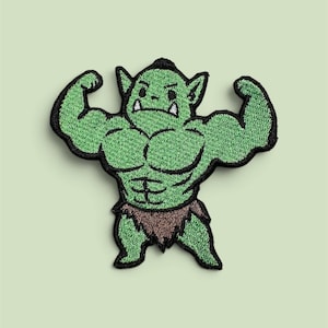 Puede incluir: Un parche bordado que muestra un personaje musculoso de color verde con orejas puntiagudas y dos colmillos blancos. El personaje flexiona ambos brazos, mostrando músculos pectorales y abdominales definidos, y viste un taparrabos marrón con bordes negros.