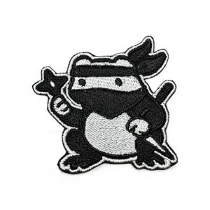 May include: Black and white embroidered patch of a ninja frog. The frog is depicted with a ninja mask and headband, holding a shuriken and a katana. The patch has a white border.