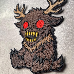 Puede incluir: Parche bordado con una criatura de dibujos animados con pelaje marr&oacute;n, cuernos y dientes amarillos. La criatura tiene grandes ojos rojos y garras negras. El parche est&aacute; dise&ntilde;ado para planchar o coser sobre tela.