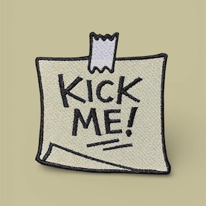 May include: An embroidered patch shaped like a square sticky note in off-white with a black border. The text KICK ME! is stitched in black letters in the centre. A small piece of white tape is stitched at the top, and one corner is folded up.