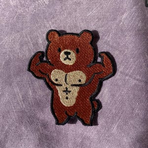 Puede incluir: Parche bordado con un oso pardo musculoso flexionando sus brazos. El oso tiene un pecho beige y un contorno negro. Ideal para ropa o accesorios.