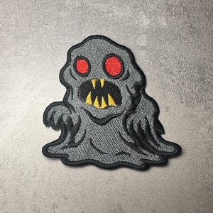 Pode incluir: Patch bordado com um monstro cinzento amorfo com olhos vermelhos e uma boca sorridente. O monstro tem um contorno preto e está sobre um fundo cinzento. O patch é projetado para roupas ou acessórios.
