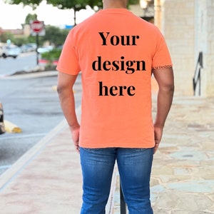 May include: A person wearing a bright orange t-shirt with the text "Your design here" printed on the back. The shirt is a blank canvas for customization.