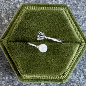 May include: A silver ring with a clear, round gemstone and a silver ring with a white, round gemstone, displayed in a green velvet ring box. The ring box is hexagonal.